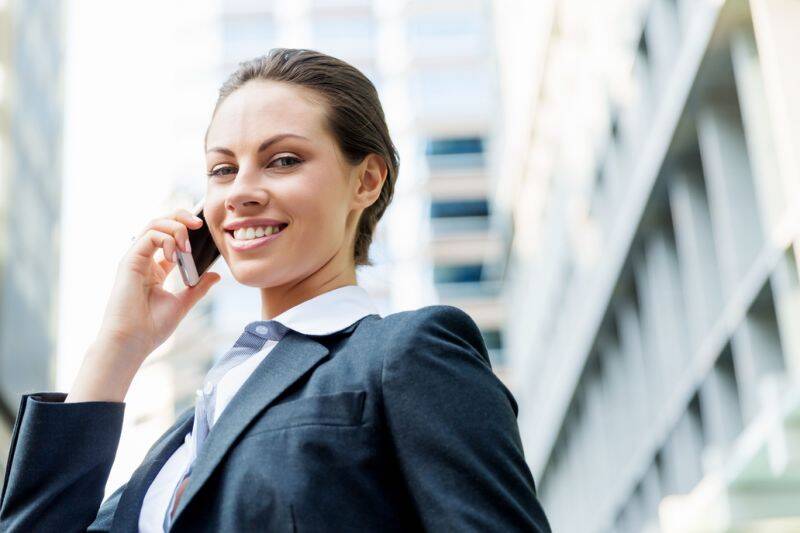 A smiling businesswoman in a suit talking on the phone outside a modern office building.