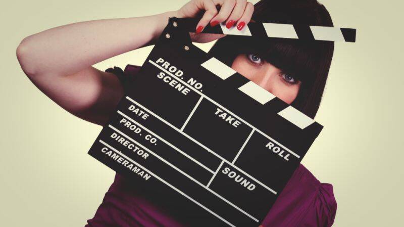Eine Frau in einem violetten Kleid hält eine Filmklappe, die die Finanzplanung und die Vermögensverwaltungsstrategien von Prominenten symbolisiert.